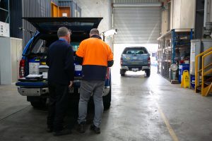 A Tandex technician and factory hand review paperwork at the back of a blue service vehicle inside the warehouse, ensuring chemicals are correctly loaded for delivery to customer sites. Another Tandex vehicle is visible in the background near the roller door.