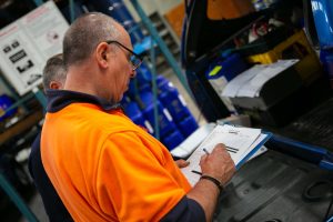 A Tandex technician and factory hand in high-visibility clothing review a printed checklist on a clipboard beside a service vehicle in the factory, with blue chemical drums and equipment visible in the background.