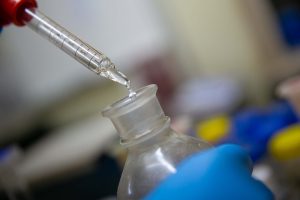 Close-up of a laboratory pipette releasing a drop of liquid into the opening of a glass sample bottle, held steady by a gloved hand.
