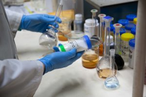 A Tandex laboratory technician wearing gloves and a lab coat handles a labelled test container and a glass flask filled with clear liquid on a workbench with various other beakers and chemical samples.
