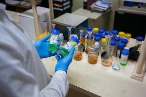 A Tandex laboratory technician wearing gloves and a lab coat holds two labelled sample vials containing green chemical solutions, with various other samples and glassware on the laboratory bench.