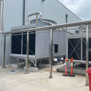 Two industrial cooling towers installed side by side outdoors, with the larger unit on the left and a smaller tower on the right, connected by stainless steel pipework on a concrete pad.