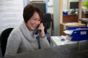 A Tandex office staff member smiling while speaking with a customer on the phone at her desk, surrounded by office paperwork and folders, providing friendly assistance.