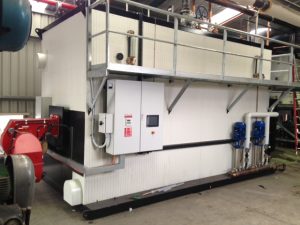 Large industrial boiler installed on a customer site, featuring a red burner assembly, control panel, circulation pumps, and overhead pipework with an access platform above the unit.