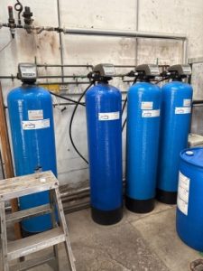 Multiple blue water softener units connected by piping and control valves at a customer facility.