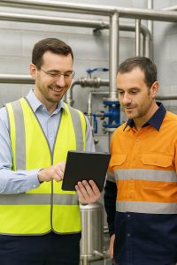 An engineer wearing a high-visibility vest and a plant room technician in an orange and navy uniform review operational data on a tablet inside a plant room surrounded by stainless steel piping and industrial equipment.